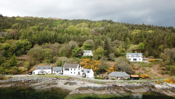 Un village côtier aux maisons blanches, avec un littoral rocheux et, en arrière-plan, des collines densément boisées.