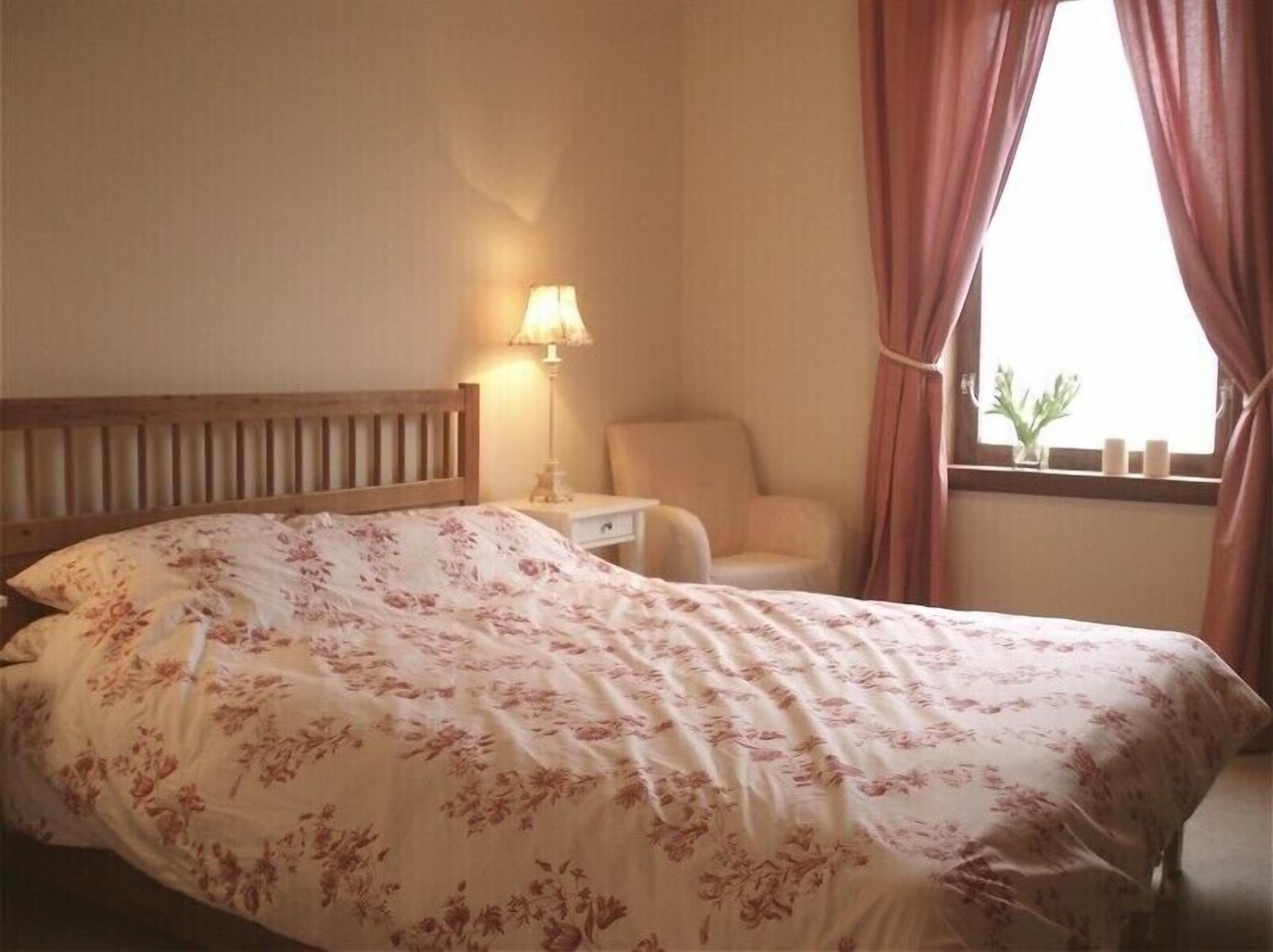 A bedroom with a bed, a nightstand, a lamp, a chair, and a window with curtains.