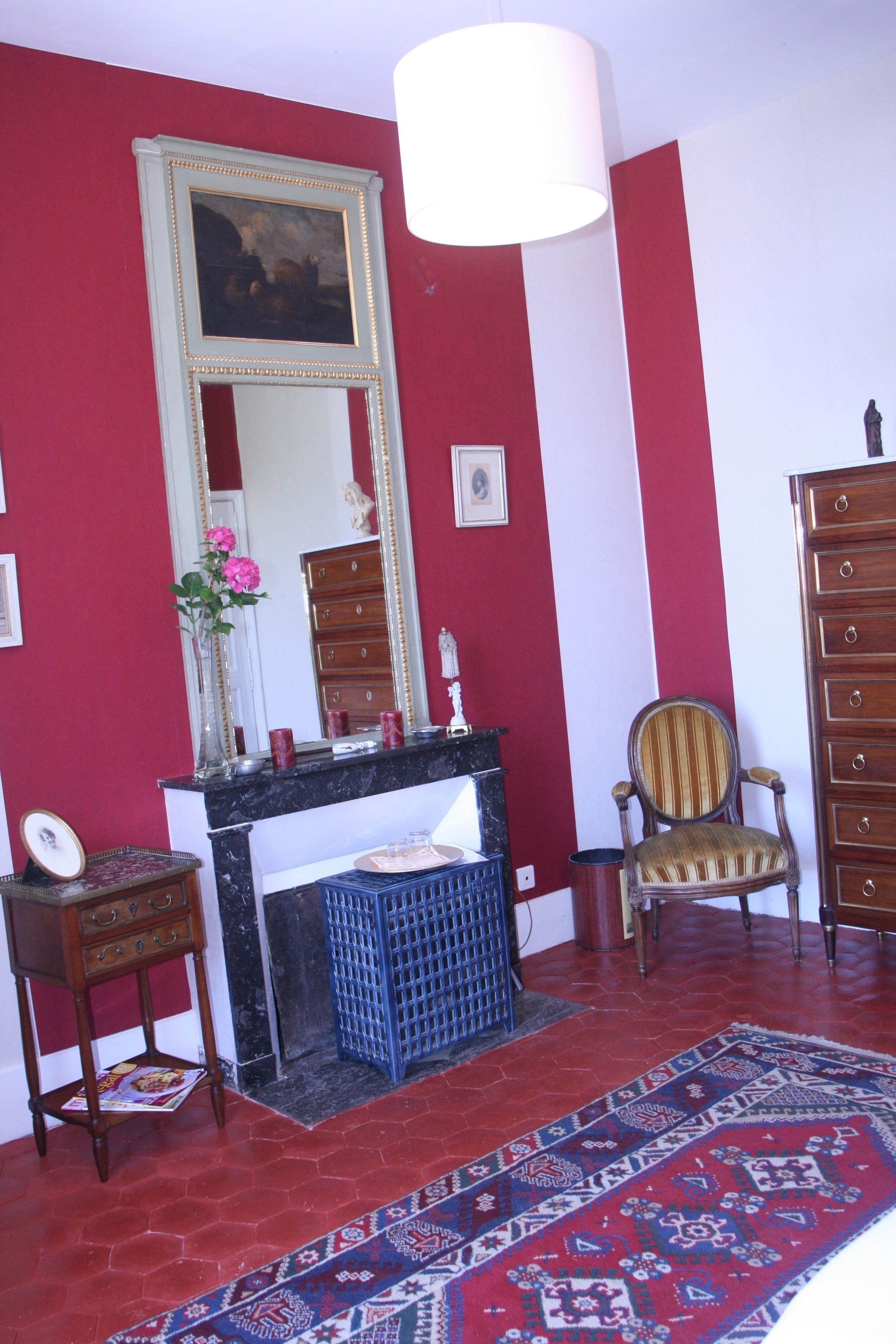 A room with a red and white wall, a large mirror, a wooden dresser, a chair, a small table with a vase of flowers, and a patterned rug.