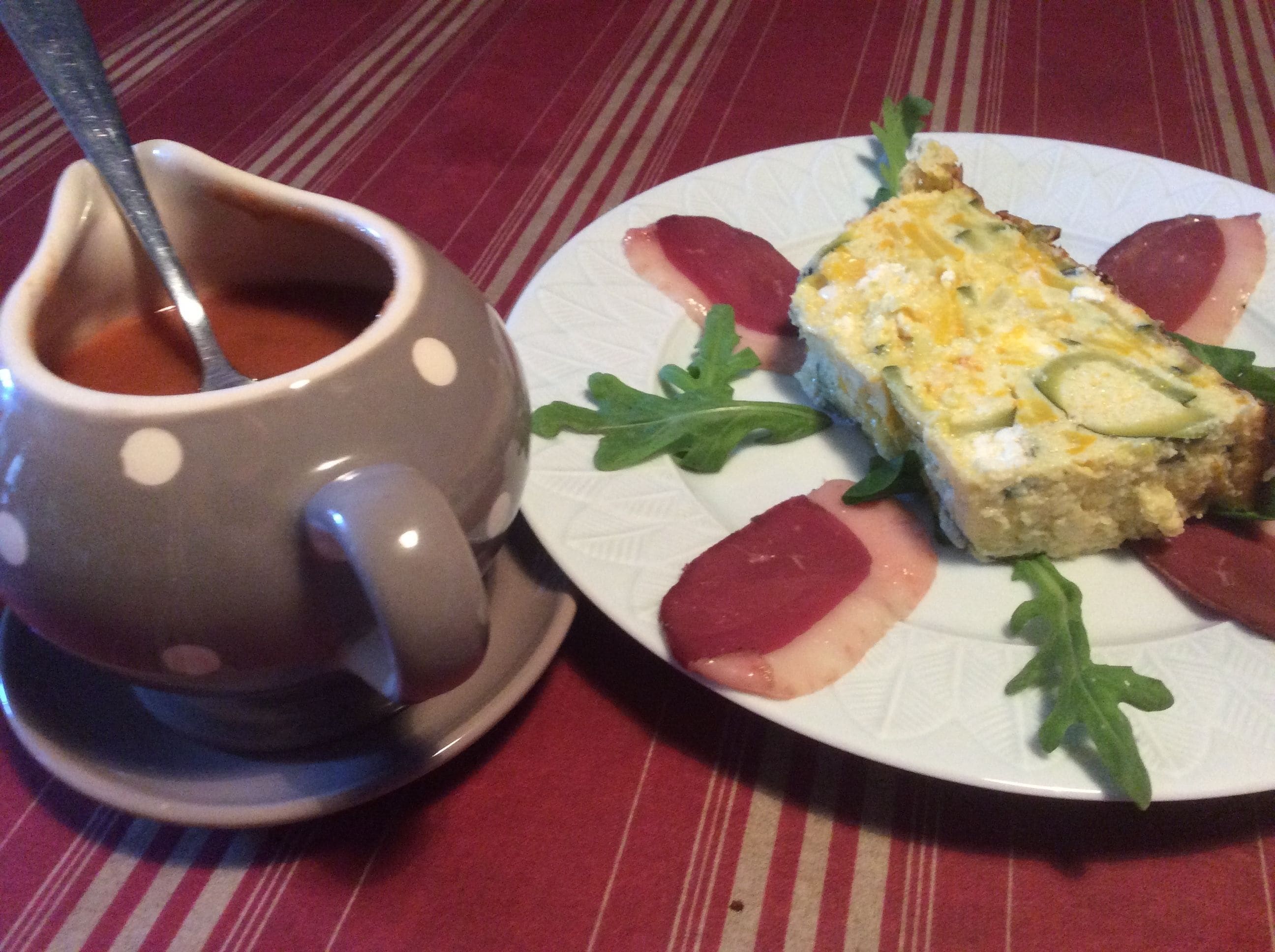 A plate with a slice of quiche and radish slices, a cup with a spoon, and a red and white striped tablecloth.