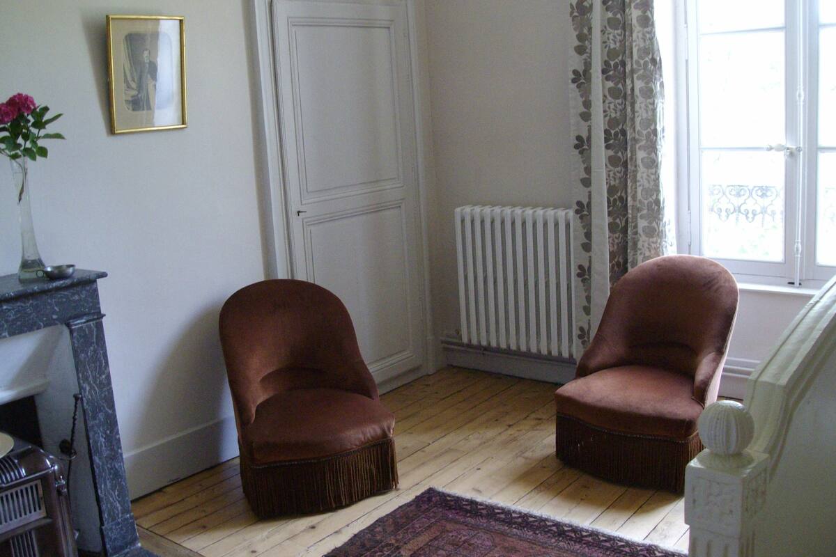 Une pièce avec deux fauteuils marron, un radiateur, une porte blanche et une fenêtre ornée de rideaux à motifs floraux.