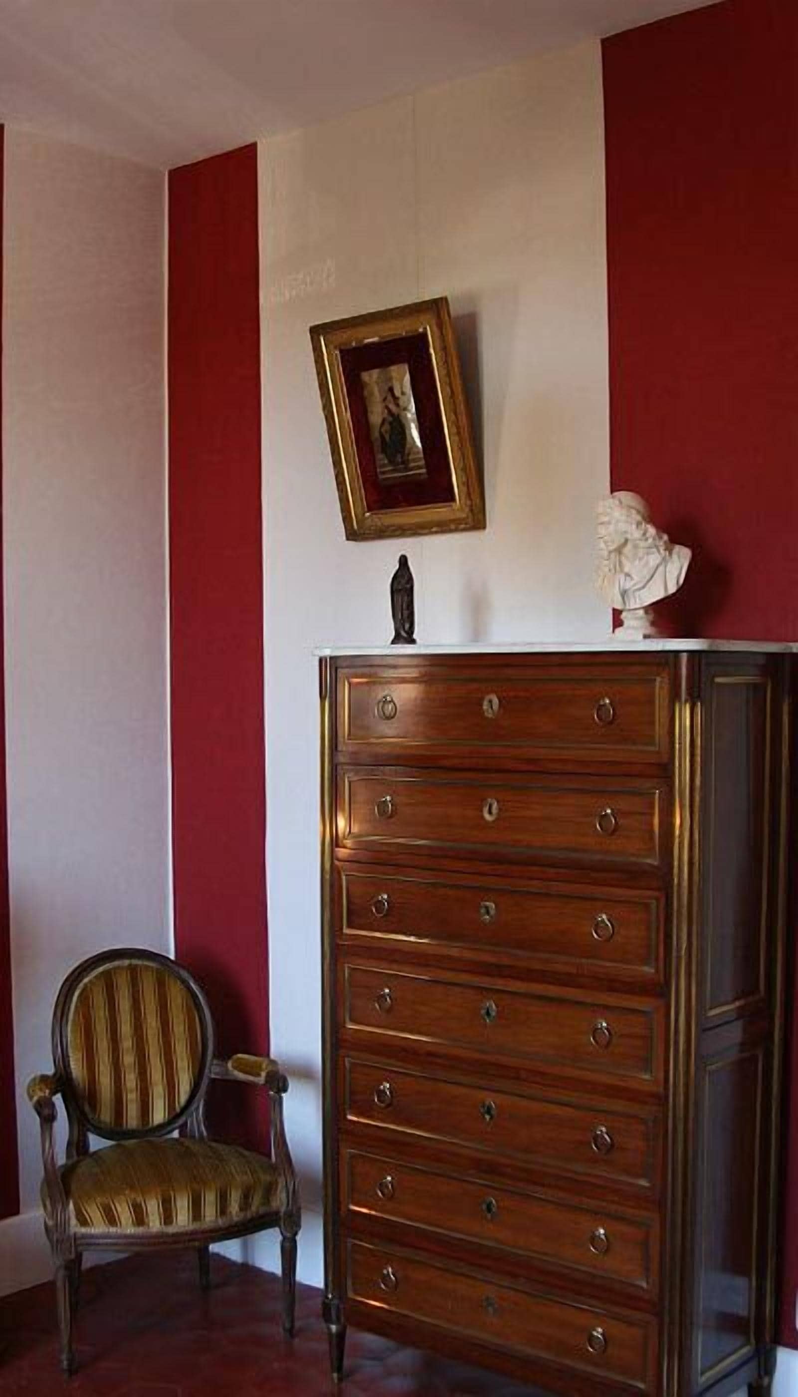 A wooden dresser with multiple drawers and a framed picture on top, a chair with a striped cushion, and red and white walls.