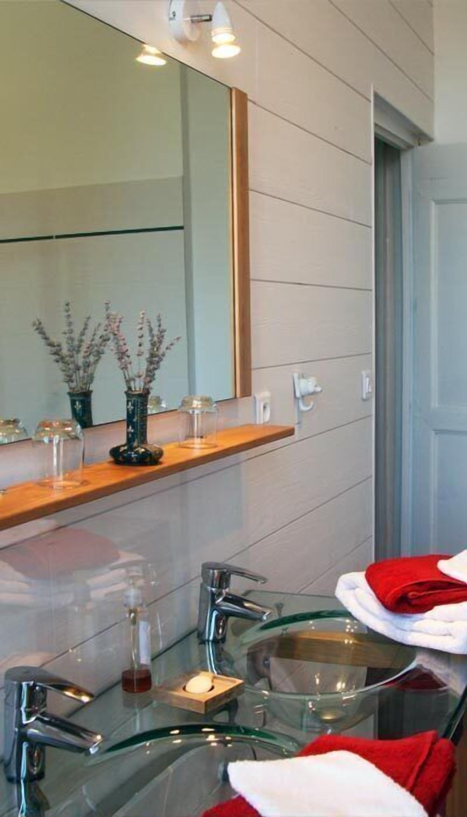 Une salle de bain moderne avec un lavabo en verre, un grand miroir et une étagère en bois agrémentée d’objets décoratifs.