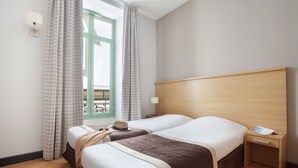 A hotel room with two beds, a wooden headboard, a nightstand, a window with curtains, and a view of the outdoors. - Appartment sympa 4p, près des musées et de la gare (Nice)