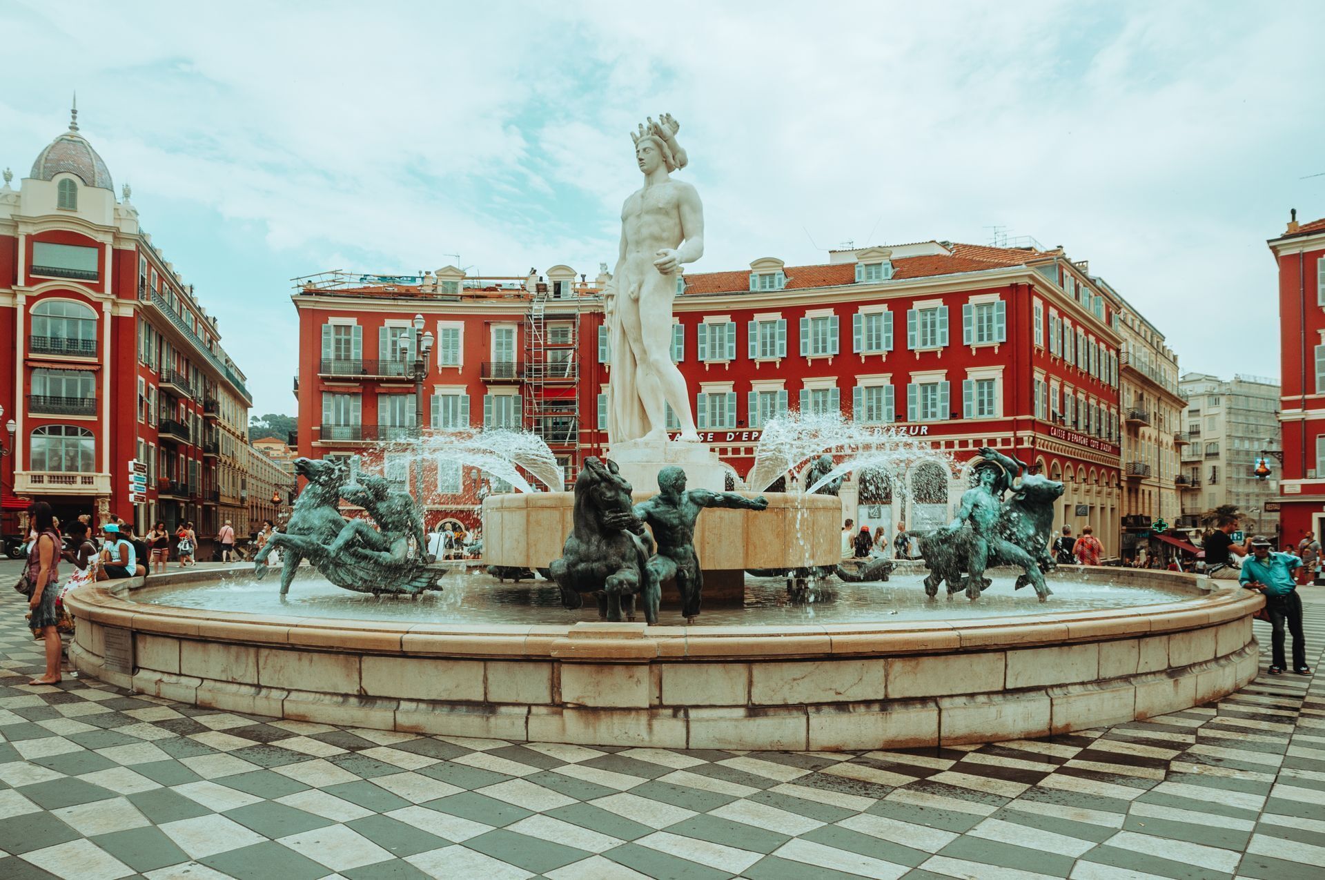 Ein öffentlicher Platz mit einem zentralen Brunnen, der Statuen und Wasserfontänen zeigt, umgeben von roten Gebäuden mit Balkonen.