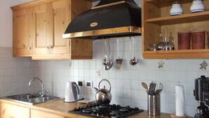 Een keuken met houten kastjes, een gasfornuis, een waterkoker en hangend keukengerei.