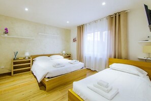A hotel room with two beds, wooden flooring, a small shelf with decorative items, and a window with curtains. - Welc-om Vittoria (Padova)