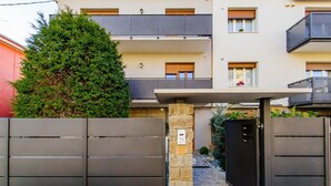 Un immeuble d’appartements moderne, doté d’une entrée sécurisée par un portail, d’un pilier en pierre et d’un grand arbre à feuilles persistantes.