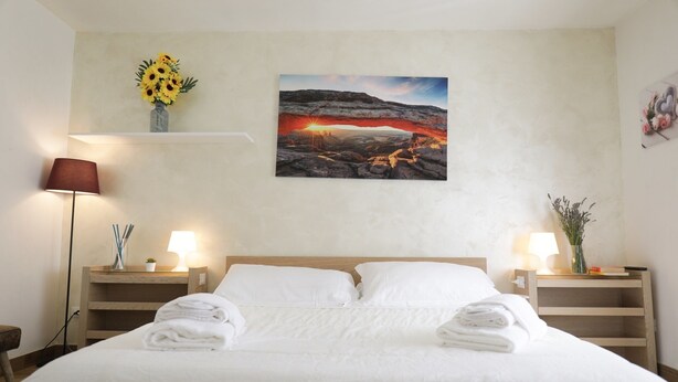 Un lit bien fait, avec du linge de lit et des serviettes blancs, deux lampes de chevet, un tableau représentant un coucher de soleil sur un paysage rocheux, et un vase rempli de fleurs jaunes, posé sur une étagère au-dessus du lit.