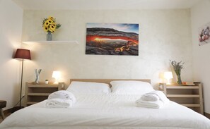 A neatly made bed with white linens and towels, two bedside lamps, a painting of a sunset over a rocky landscape, and a vase with yellow flowers on a shelf above the bed. - Welc-om Vittoria (Padova)