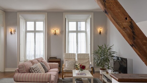 A living room with a sofa, armchairs, a coffee table, and a TV. There are two windows with curtains, a wooden beam, and a plant.