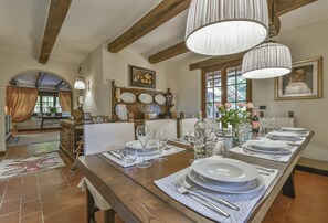Uma área de jantar com uma longa mesa de madeira posta para uma refeição, cercada por cadeiras. Há pratos, copos e talheres sobre a mesa. O espaço possui vigas de madeira aparentes no teto e uma grande janela com cortinas.