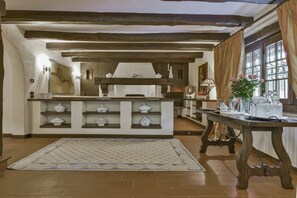 Uma cozinha rústica com vigas de madeira aparentes, uma ilha central com armários brancos e uma grande janela com cortinas.