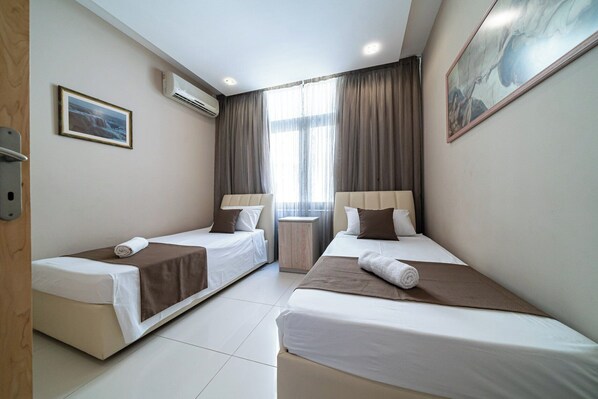 A hotel room with two single beds, a large window with curtains, an air conditioning unit, and a painting on the wall.