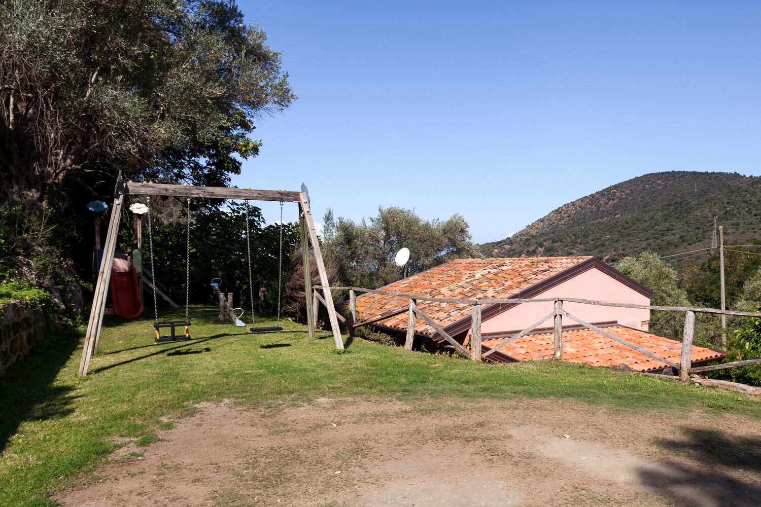 Un espace extérieur rustique avec une balançoire en bois, un toboggan rouge et un bâtiment doté d’un toit en tuiles.