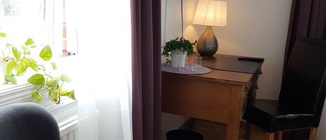 Una habitación con una silla tapizada de color negro, un escritorio de madera con una lámpara, un televisor montado en la pared y una ventana con cortinas.