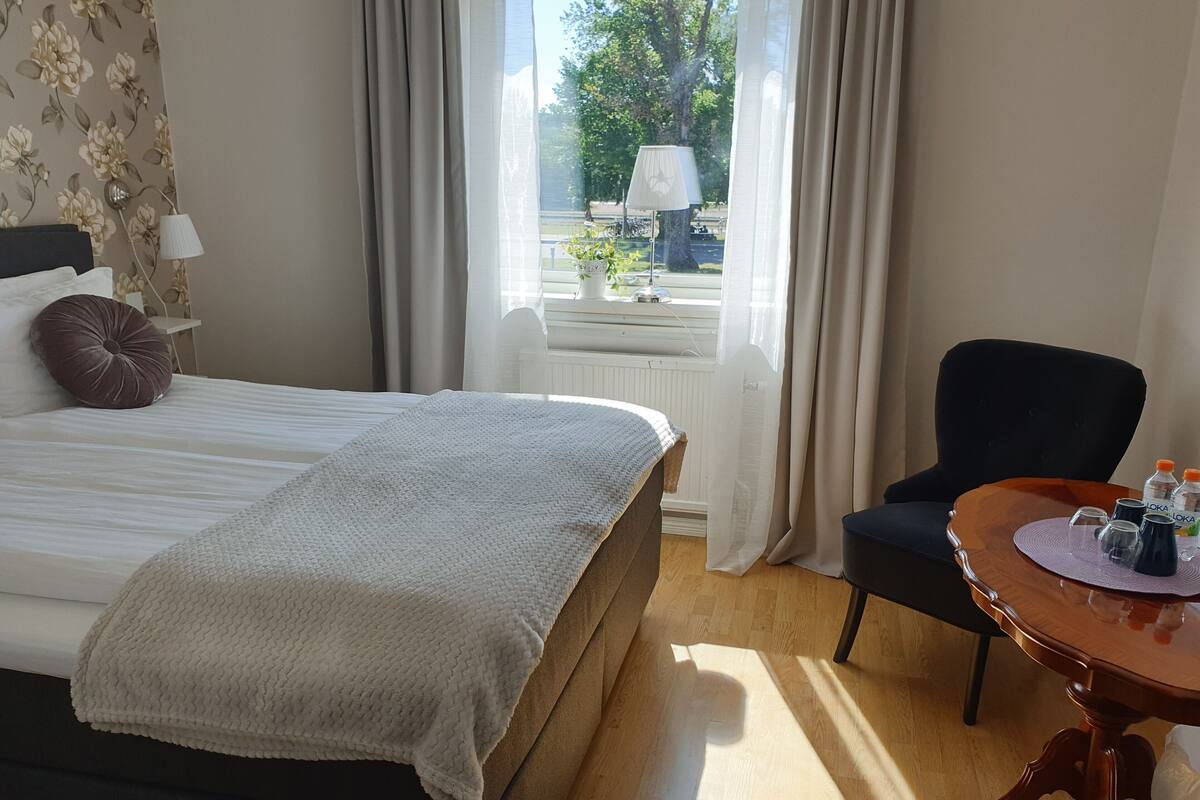 A hotel room with a bed, a chair, a small table with water bottles, and a view of the outdoors.