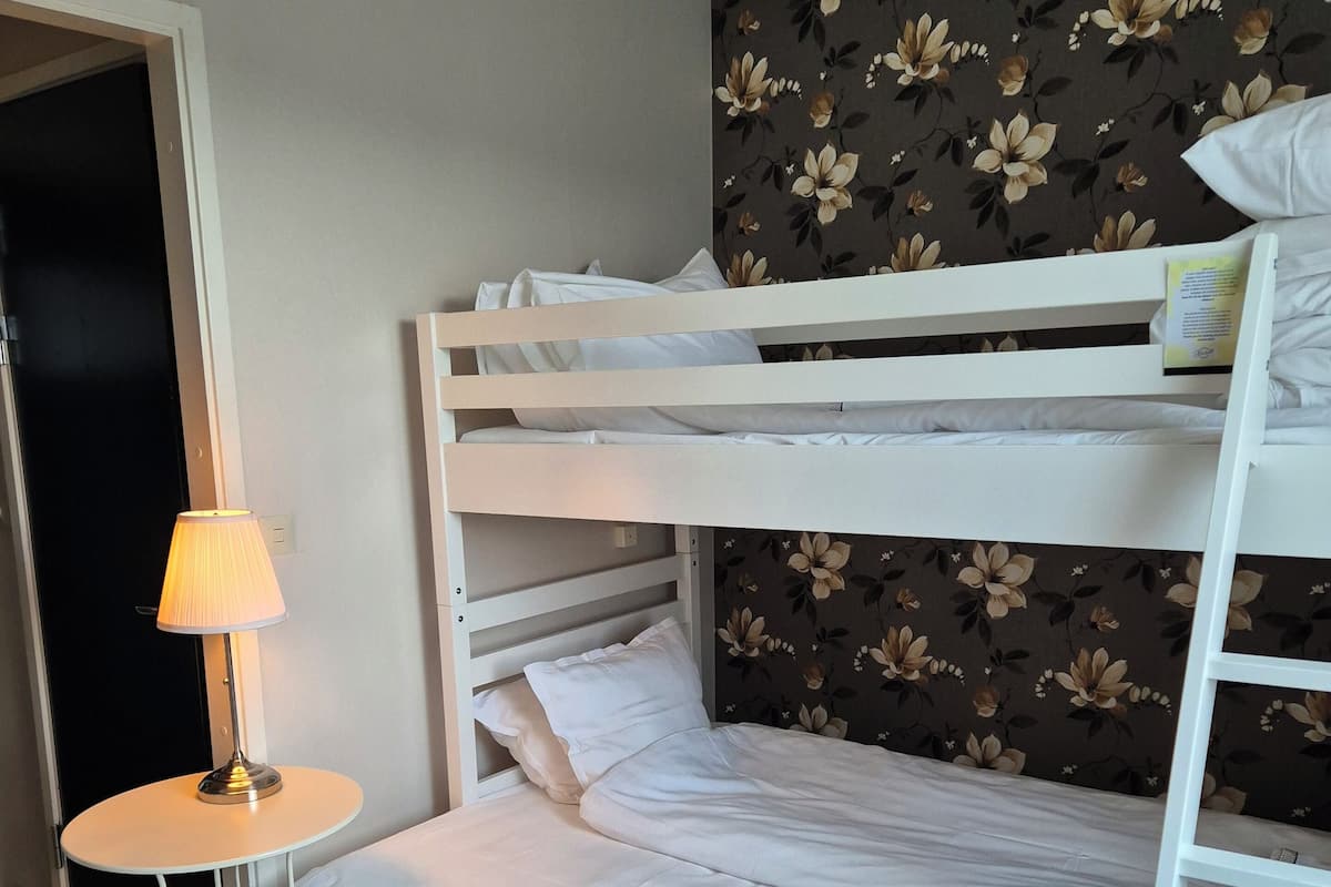 A bunk bed room with a floral wallpaper, a small round table with a lamp, and a wooden floor.