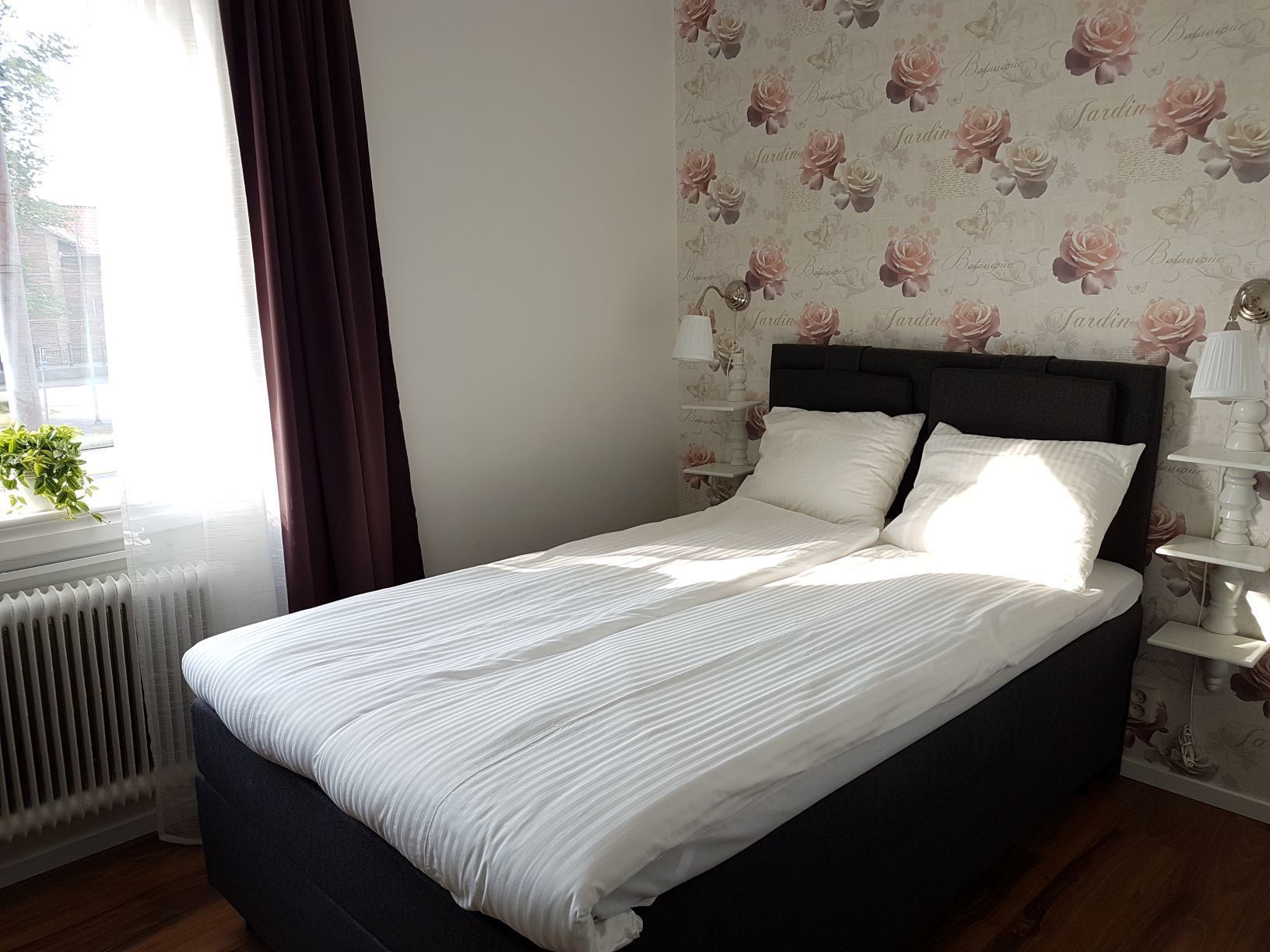 A neatly made bed with white bedding, a dark headboard, and a floral wallpaper pattern on the wall.