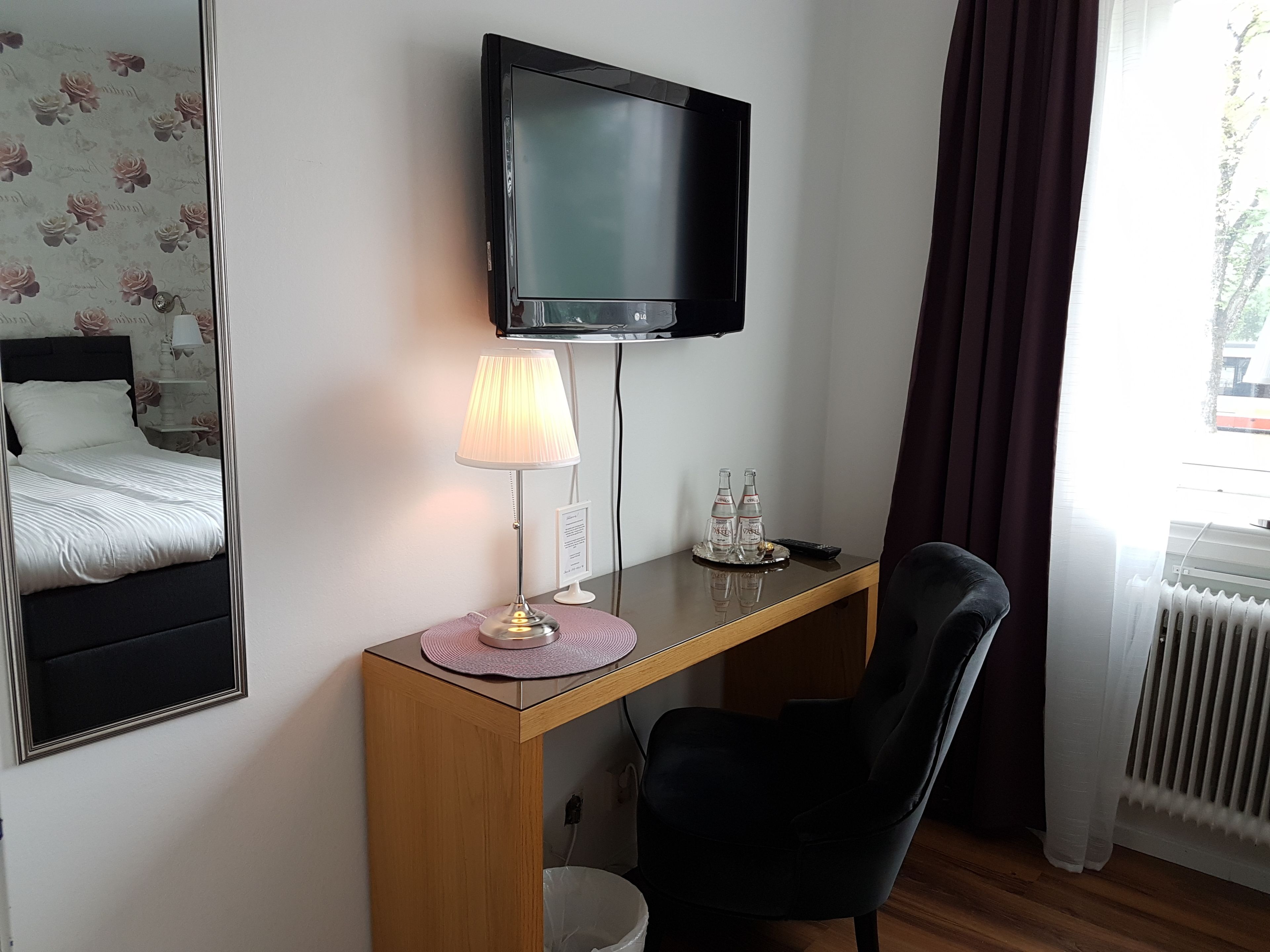 Habitación de hotel con cama, escritorio con lámpara, silla, televisión y ventana con cortinas.