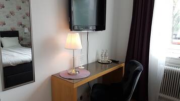 Habitación de hotel con cama, escritorio con lámpara, silla, televisión y ventana con cortinas.