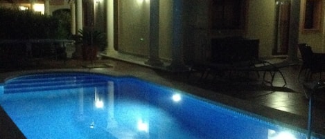 A swimming pool at night with surrounding buildings and outdoor seating.