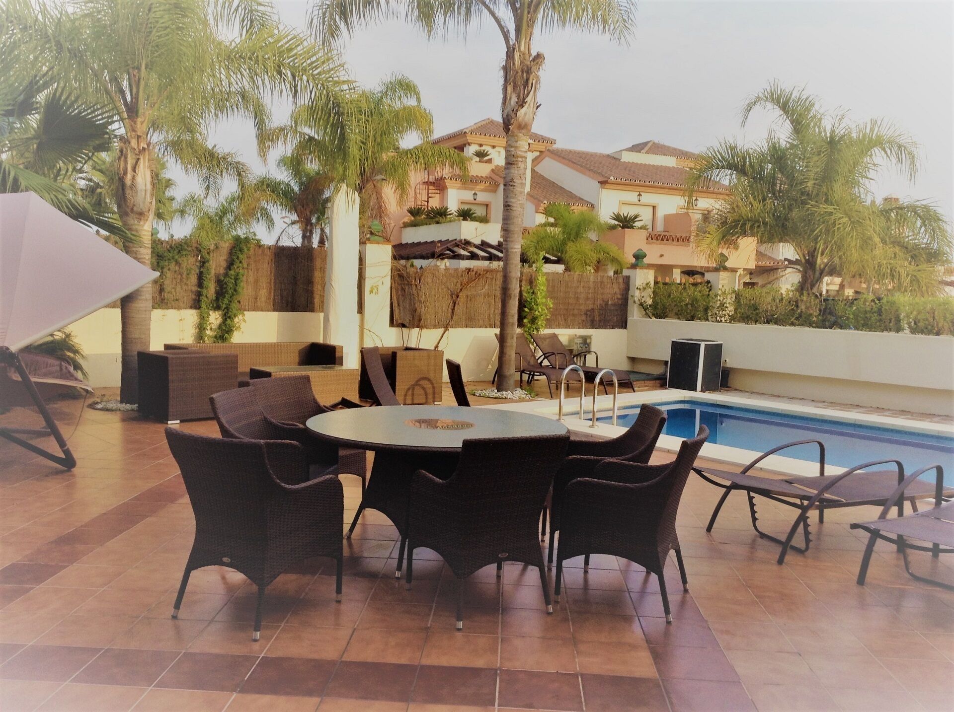 An outdoor patio with a round table and chairs, a pool, and palm trees.