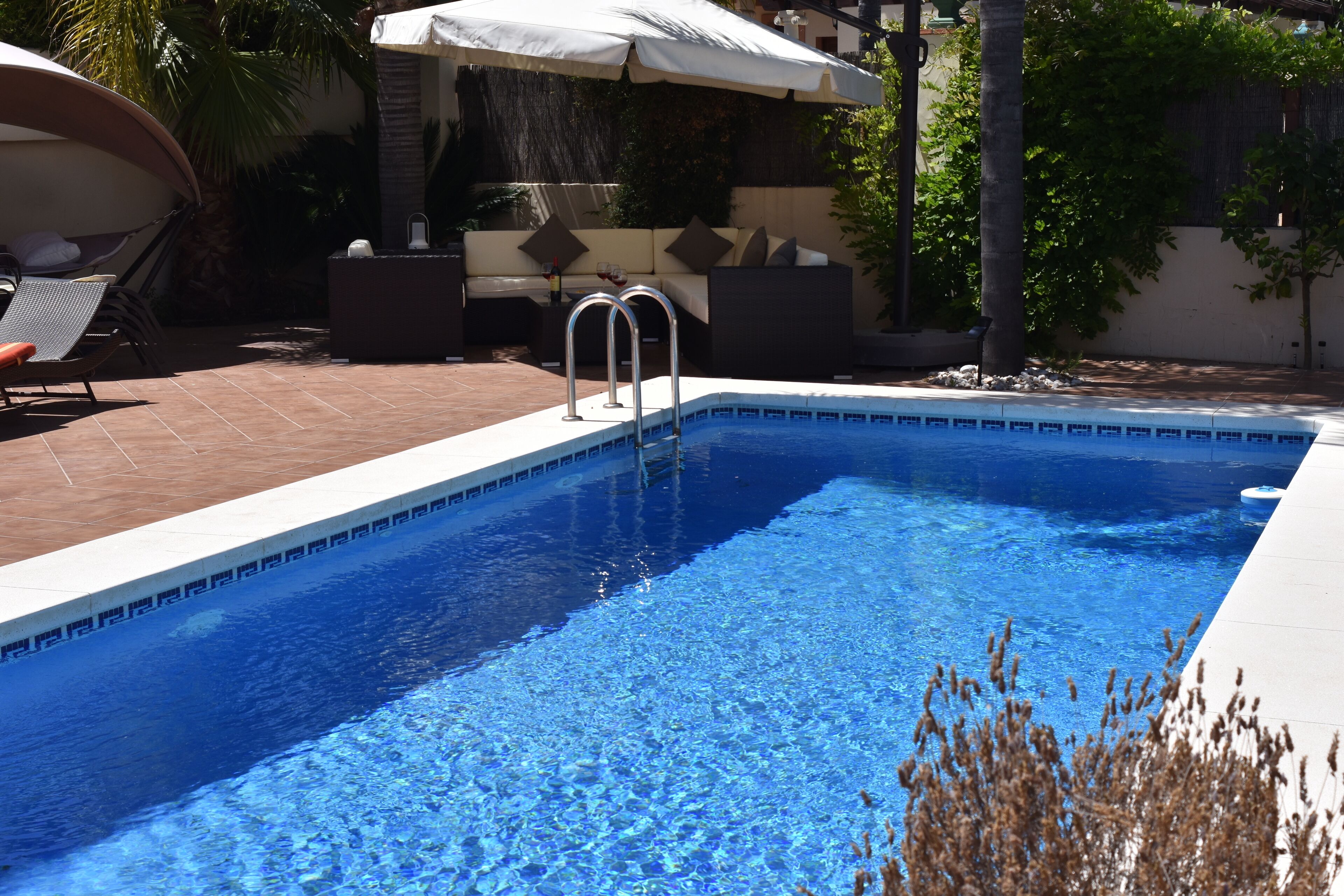 A rectangular swimming pool with a clear blue water surface, surrounded by a tiled deck. There are lounge chairs and a covered seating area with a white umbrella.