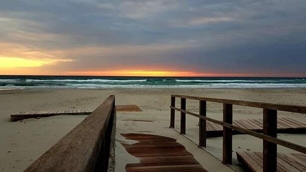 Ein hölzerner Steg führt zu einem Strand, im Hintergrund sind Wellen zu sehen.