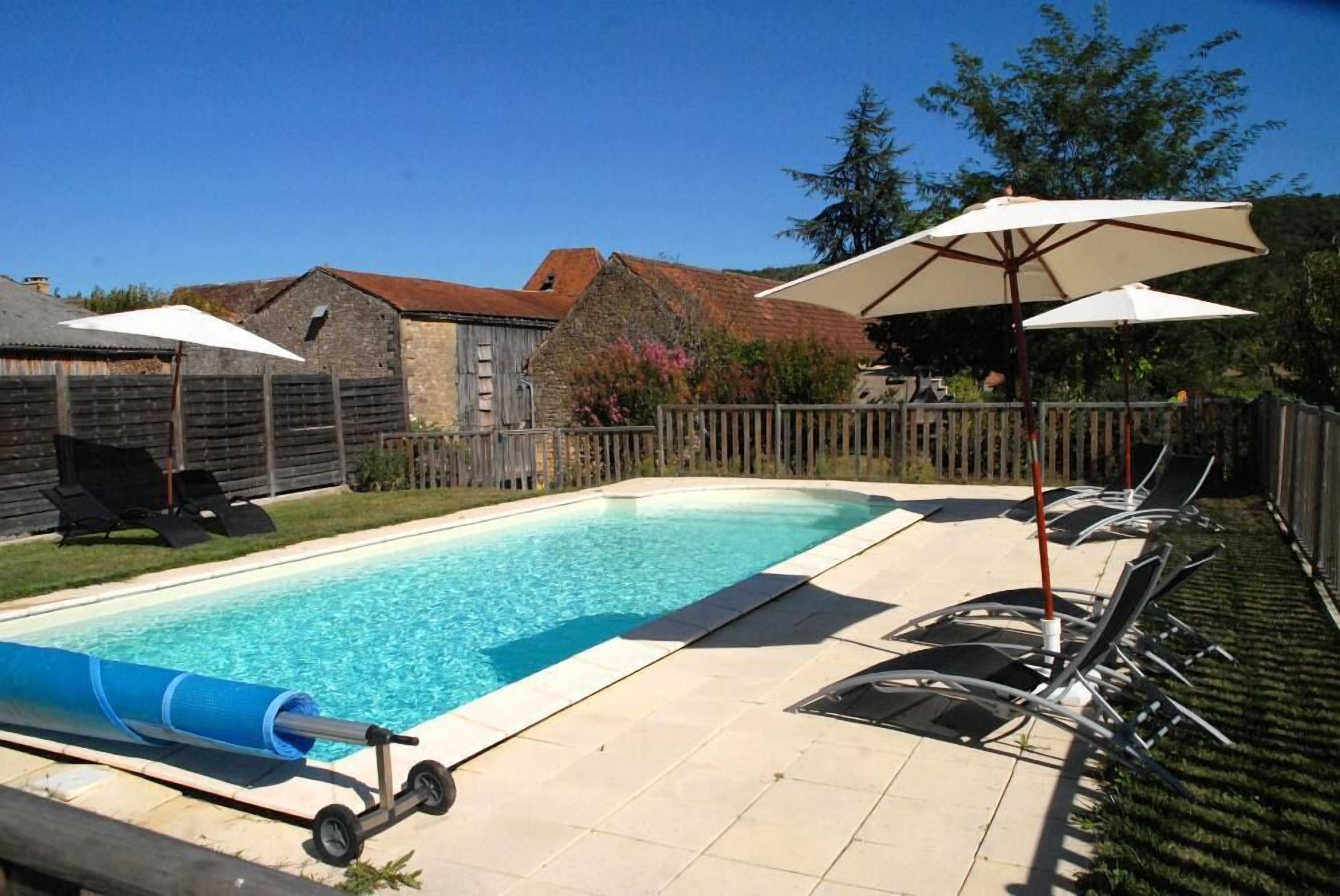 Une piscine rectangulaire avec un revêtement bleu, entourée d’une terrasse carrelée. Des chaises longues et des parasols sont disposés le long du bord de la piscine.
