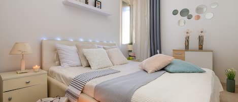 A neatly arranged bedroom with a bed, bedside tables, a nightstand, and a shelf with decorative items.