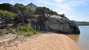 Una playa con una gran formación rocosa, un área cercada con vegetación escasa y un edificio con techo inclinado.