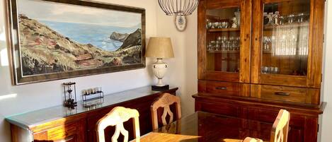 A dining area with a wooden table, chairs, a painting of a coastal landscape, a lamp, and a wooden cabinet with glass doors.