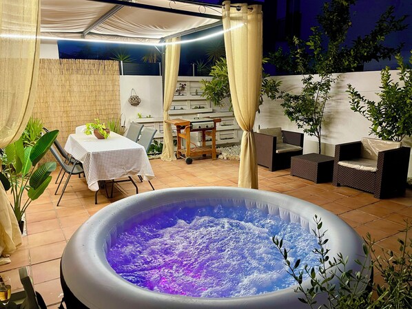 An outdoor hot tub area with a table and chairs, surrounded by potted plants and a covered seating area.