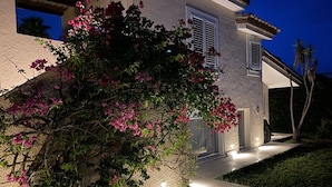 A house with a stone facade, a well-lit entrance, and a garden with pink flowers.