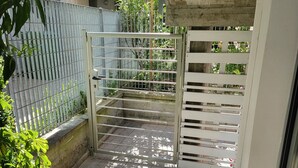 Uma área externa cercada por grades, com piso de azulejos, portão de metal e um vaso de planta.