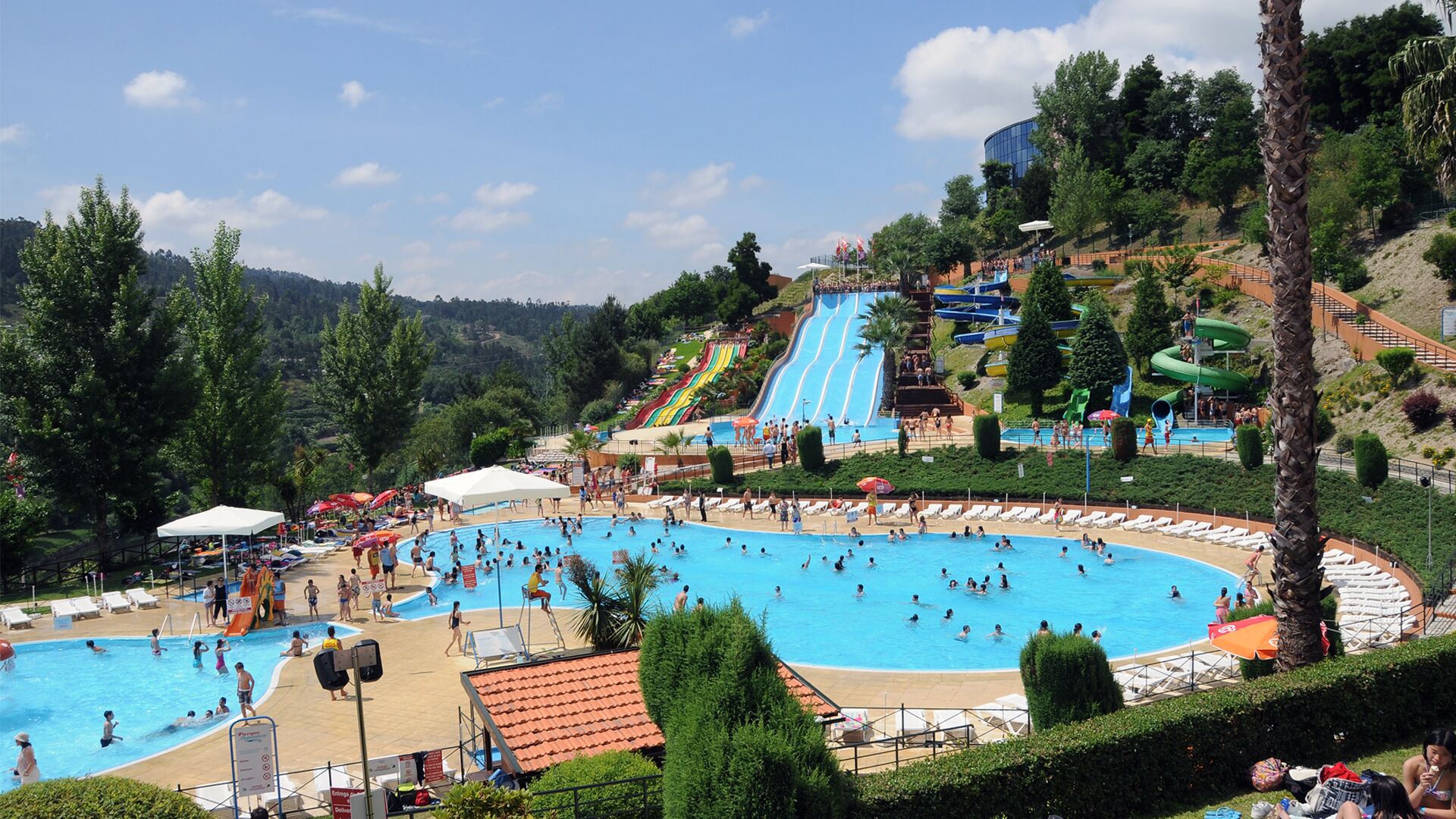 A large outdoor swimming pool surrounded by lounge chairs and umbrellas, with a water park in the background.