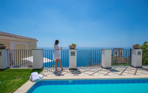 Zona de piscina con vistas despejadas al mar, una persona vestida de blanco y una villa con tejado de baldosas.