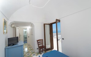 Un dormitorio con una televisión sobre un mueble azul, una silla de madera y una puerta que da a un balcón con vistas.