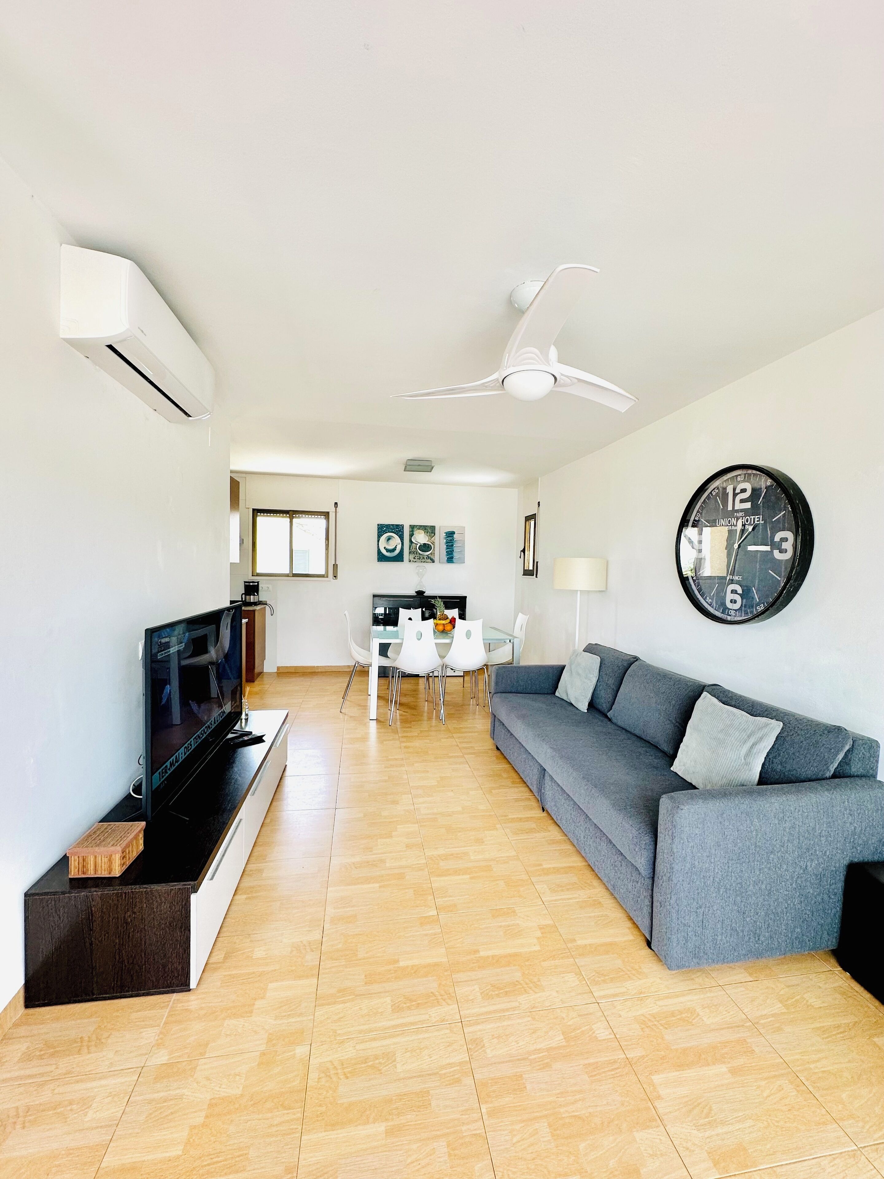 Uma sala de estar moderna com um sofá cinzento, uma televisão de tela plana e uma área de jantar com mesa e cadeiras.
