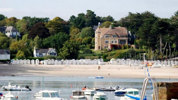 Un paysage côtier avec une plage de sable, une rangée de cabanes de plage blanches et un grand bâtiment en pierre doté d’une tour.