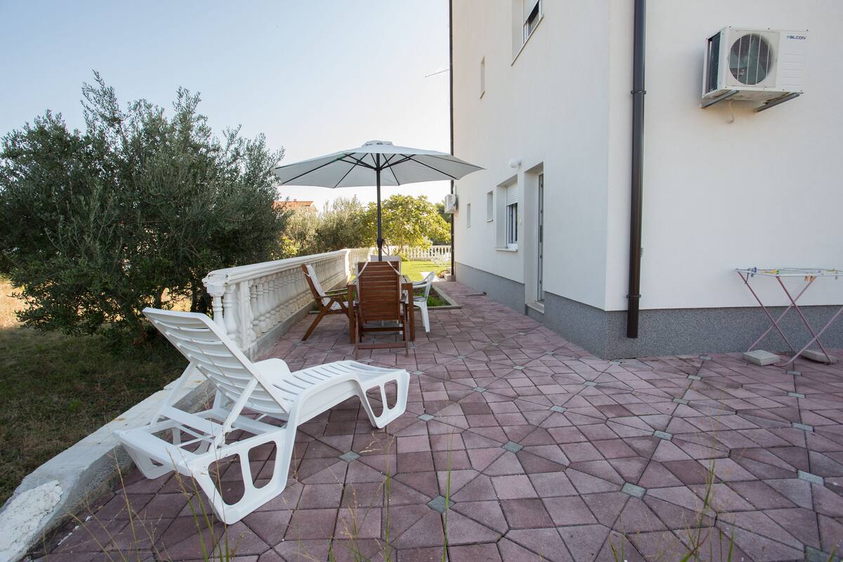 A patio with white lounge chairs and a table, surrounded by a white building with an air conditioning unit.
