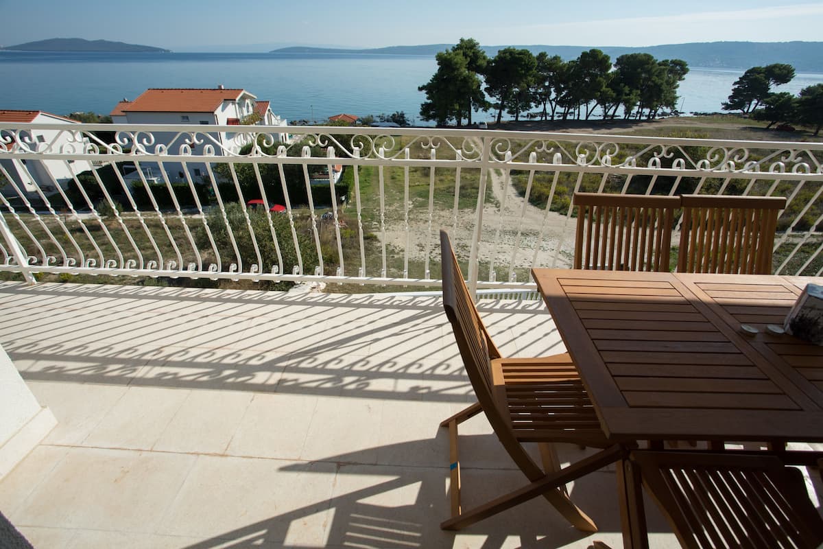 A balcony with a view of the sea and mountains, wooden furniture, and a white railing.