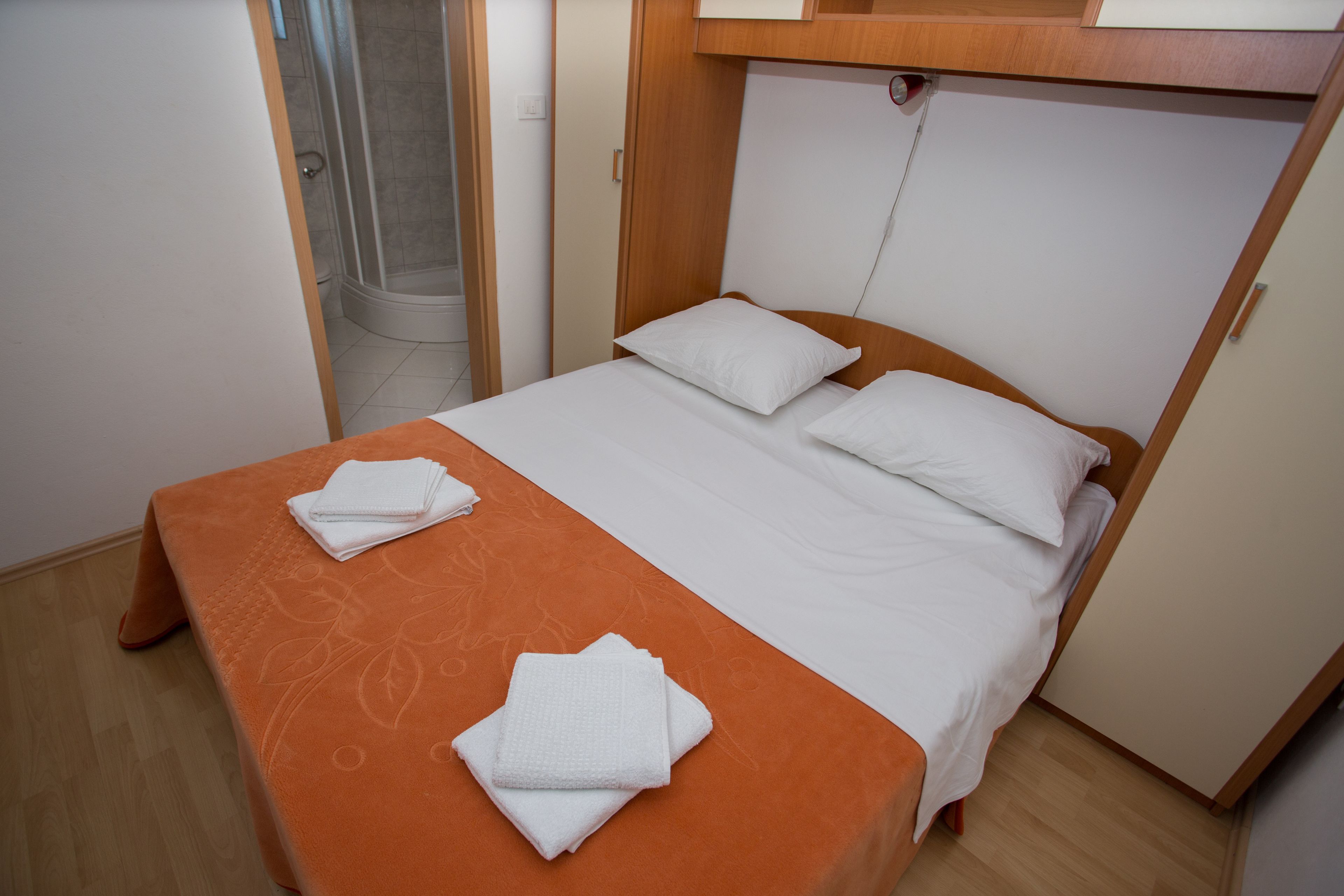 A neatly made bed with white linens and an orange bedspread, a wooden headboard, and a small wooden nightstand with a lamp.
