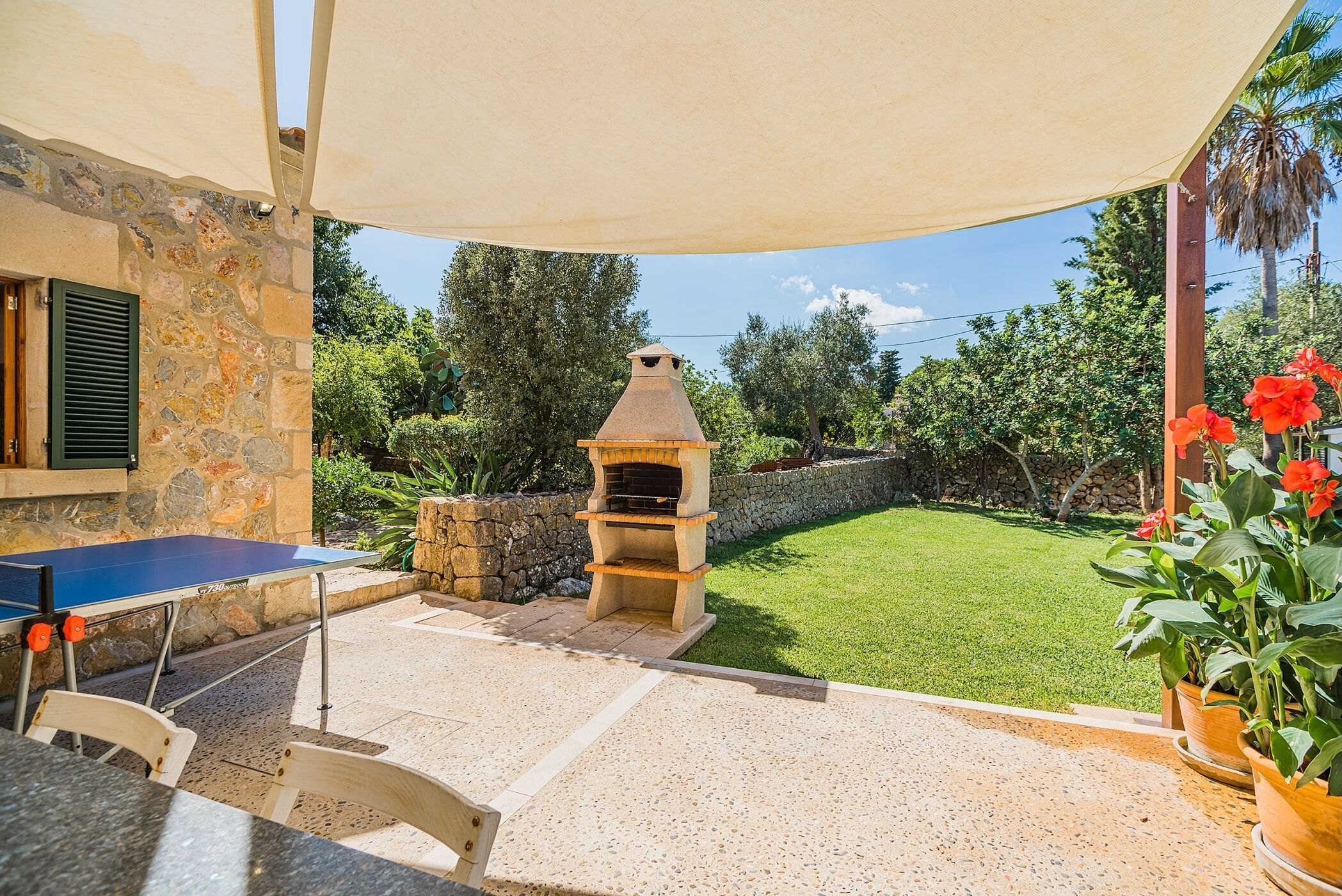 An outdoor patio with a stone wall, a barbecue grill, a ping pong table, and potted plants.