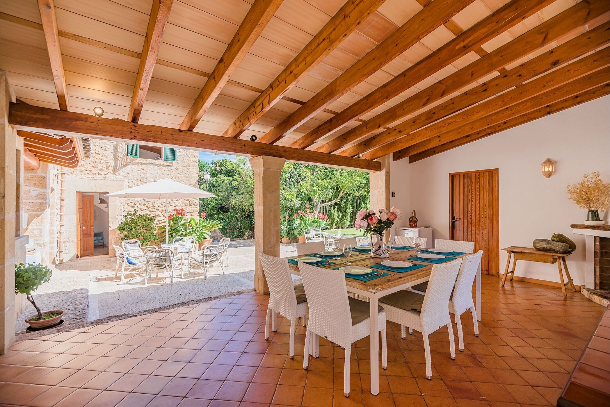 An open-plan living area with a wooden ceiling, a dining table set with white wicker chairs, and a view of the garden.
