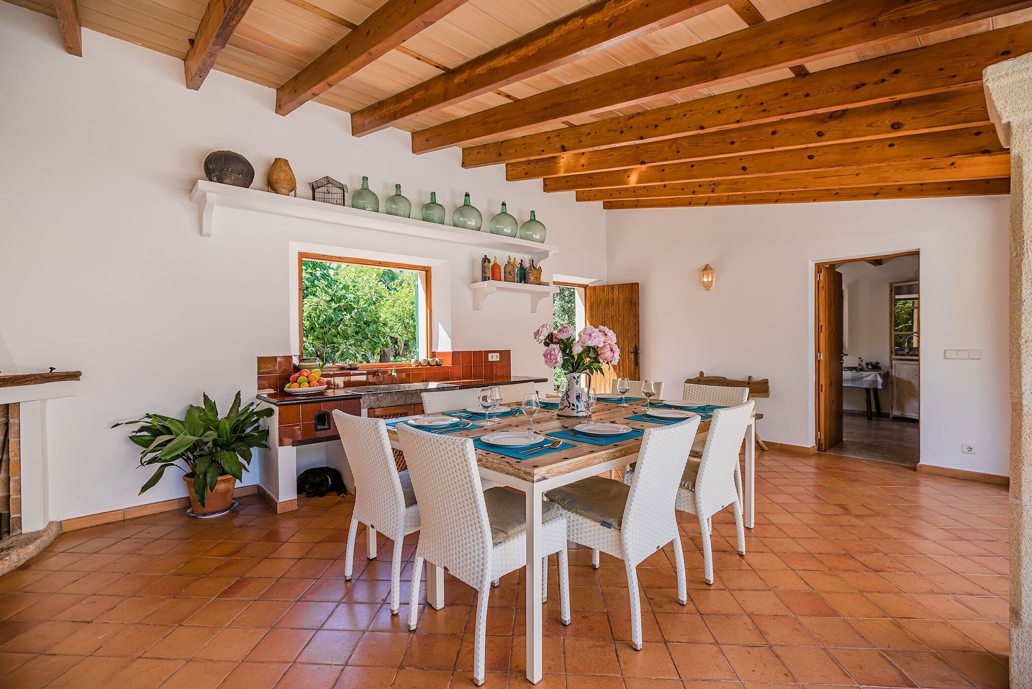 A dining area with a wooden table set for four, surrounded by white wicker chairs. A kitchen is visible in the background with a window overlooking greenery.
