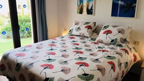 Et soveværelse med en seng, et blomstret sengetæppe, to billeder på væggen og udsigt til grøn natur gennem vinduet.