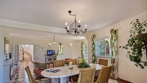 A dining area with a round table, wicker chairs, a chandelier, and a staircase leading to another room.