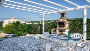 An outdoor patio with a stone-tiled floor, white plastic chairs, a table set for dining, and a barbecue grill.